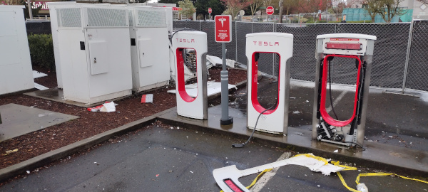 Picture taken over top of fence showing serveral tesla charging stations with destroyed section behind